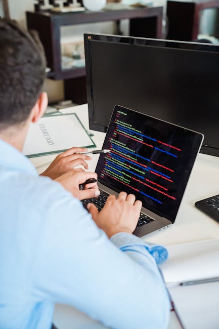 A person wearing a light blue shirt is working on a laptop at a white desk, which features a printed document, a black computer monitor, and a shelf with books and decorative items in the background.