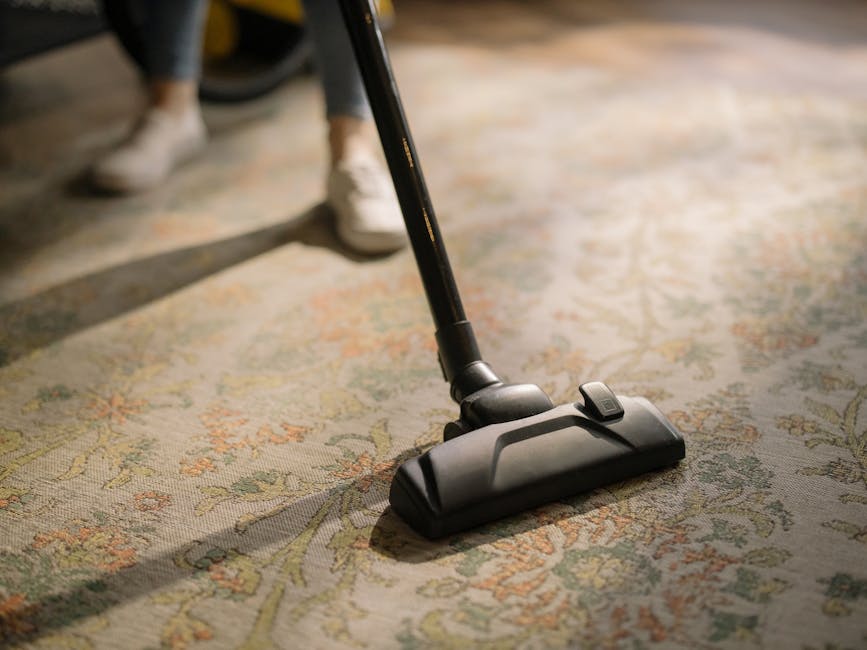 A person kneeling on a patterned carpet in a residential setting, preparing a yellow and black canister vacuum cleaner for surface cleaning. The individual is wearing a beige jacket and blue jeans, an